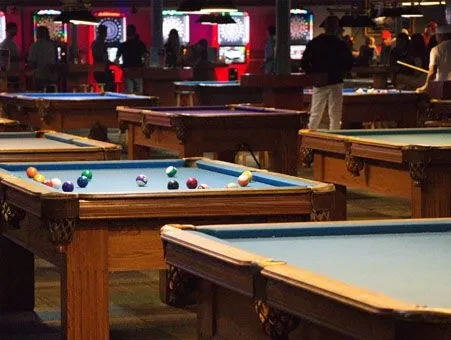 Pool hall interior with blue-felt tables, colorful balls, and people.