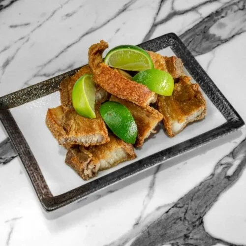Plate of crispy chicharron and lime wedges on marble.
