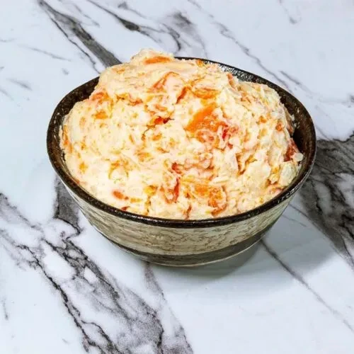 Rustic bowl of creamy, textured food with orange chunks on marble.