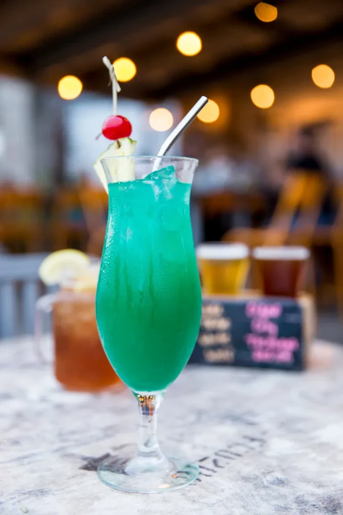 Turquoise cocktail in hurricane glass with pineapple, cherry, and straw.