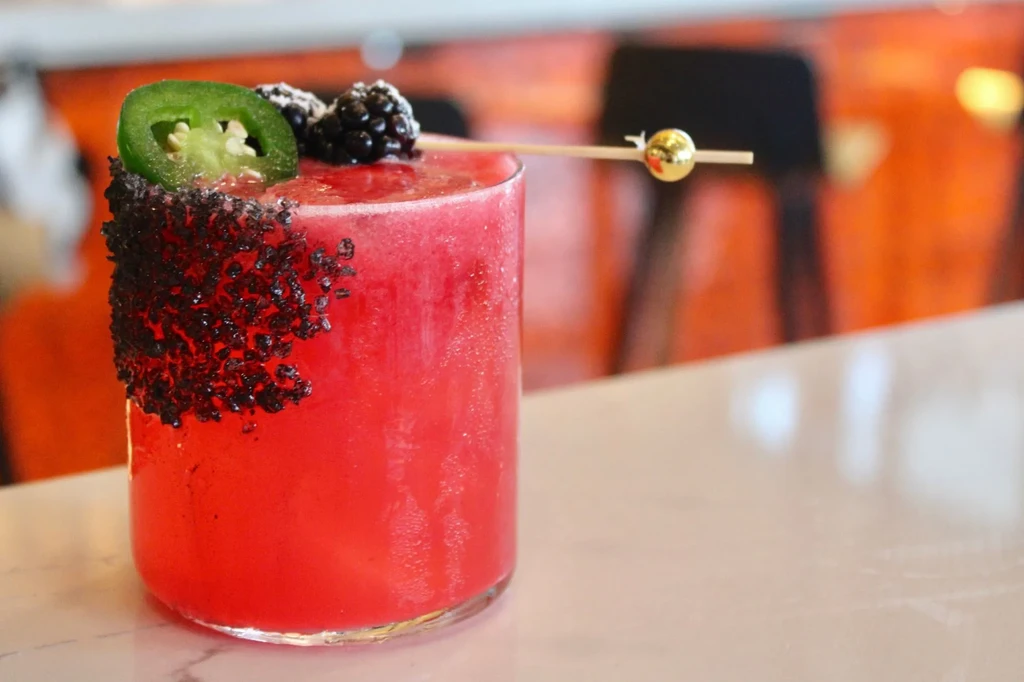 Red-pink cocktail in clear glass, garnished with jalapeño, blackberries, black rim.