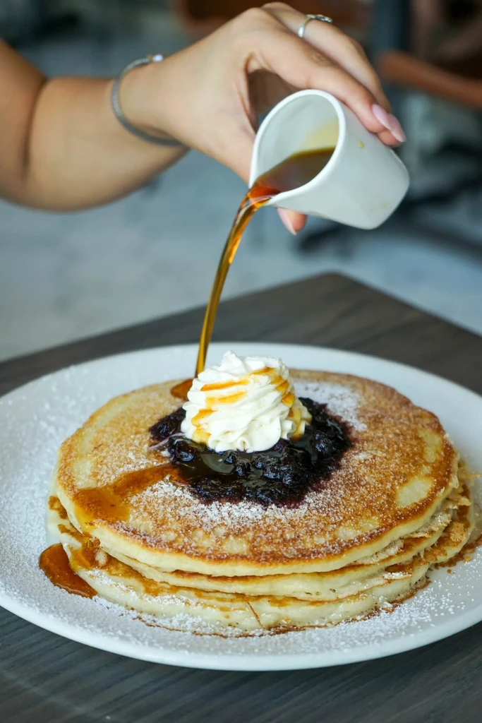 Hand pours syrup on pancakes with berries, cream, and powdered sugar.