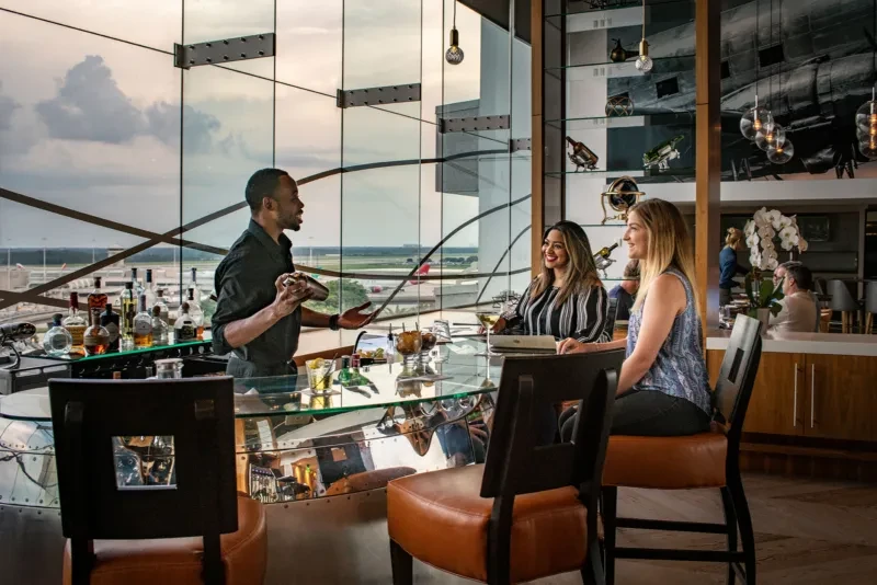 Bartender shaking drinks for two women at a bar with airport view.