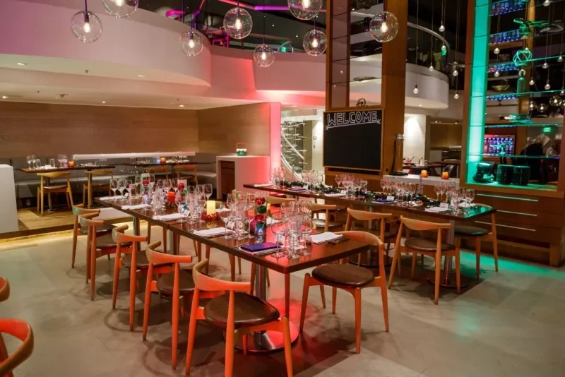 Modern restaurant interior with festive tables, vibrant lighting, and bar area.