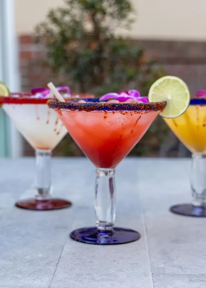 Three cocktails; central reddish-pink with seasoned rim, lime, and flower.