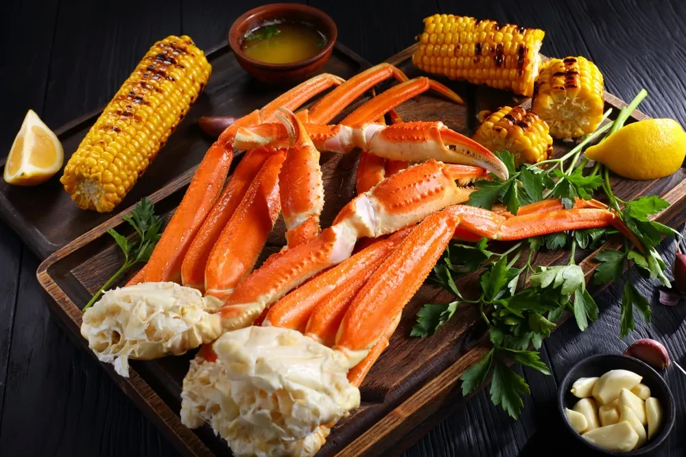 Cooked crab legs, grilled corn, lemon, and garlic on a dark board.