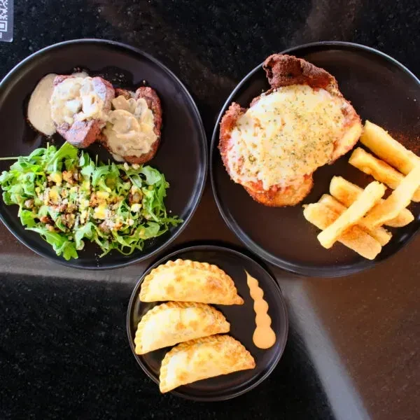 Artfully plated meat, cutlet, and empanadas on a dark table.