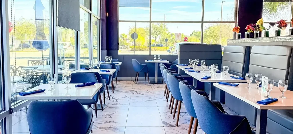 Modern restaurant dining room with set tables, blue seating, and large windows.