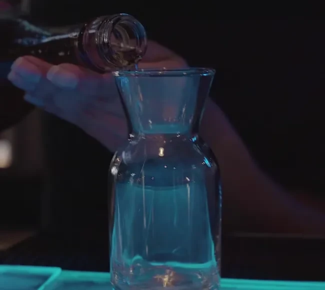 Hand pours blue-lit liquid from dark bottle into glass carafe.
