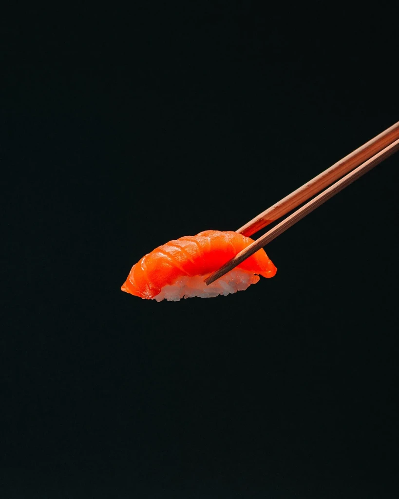 Vibrant salmon nigiri sushi held by chopsticks against dark background.