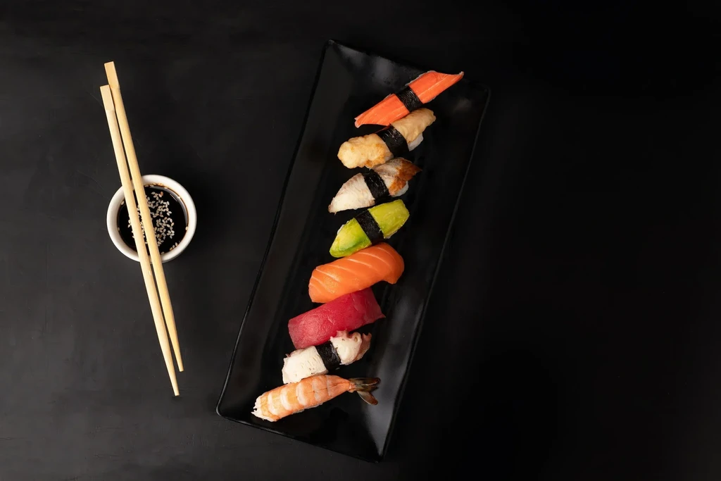 Assorted sushi nigiri, soy sauce, and chopsticks on a black plate.