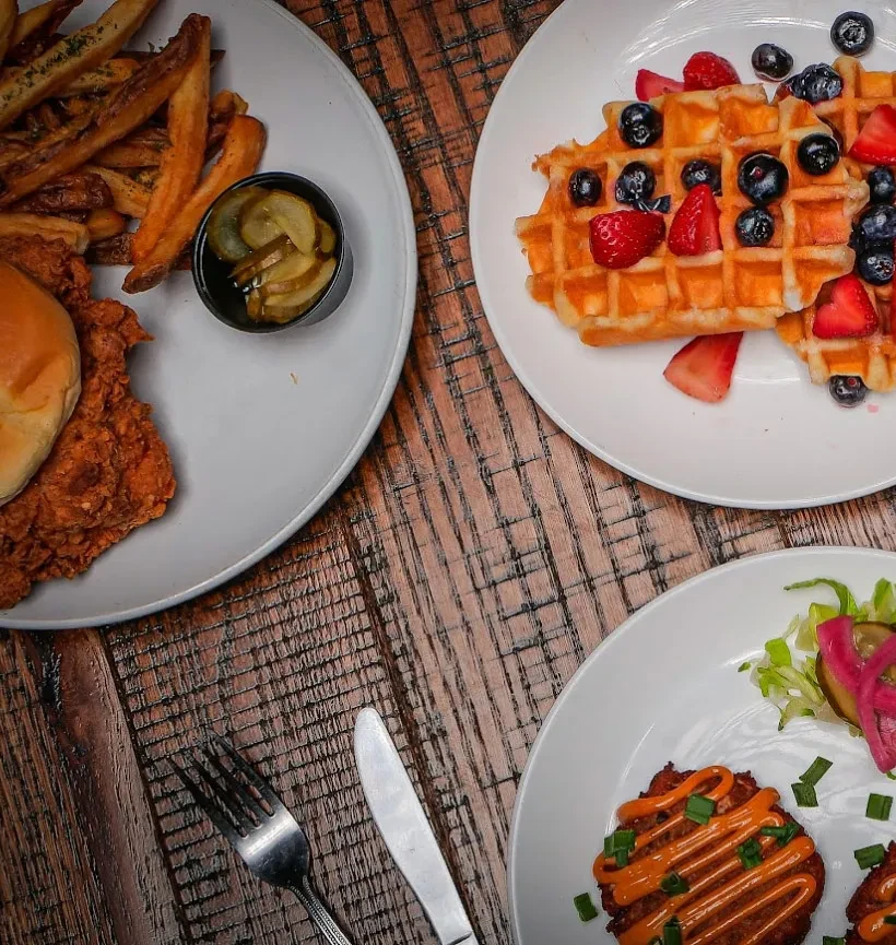 Fried chicken sandwich, waffles, and savory fritters on a wooden table.