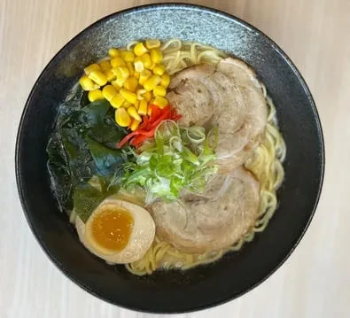 Close-up of dark ramen bowl with chashu, egg, corn, and seaweed.