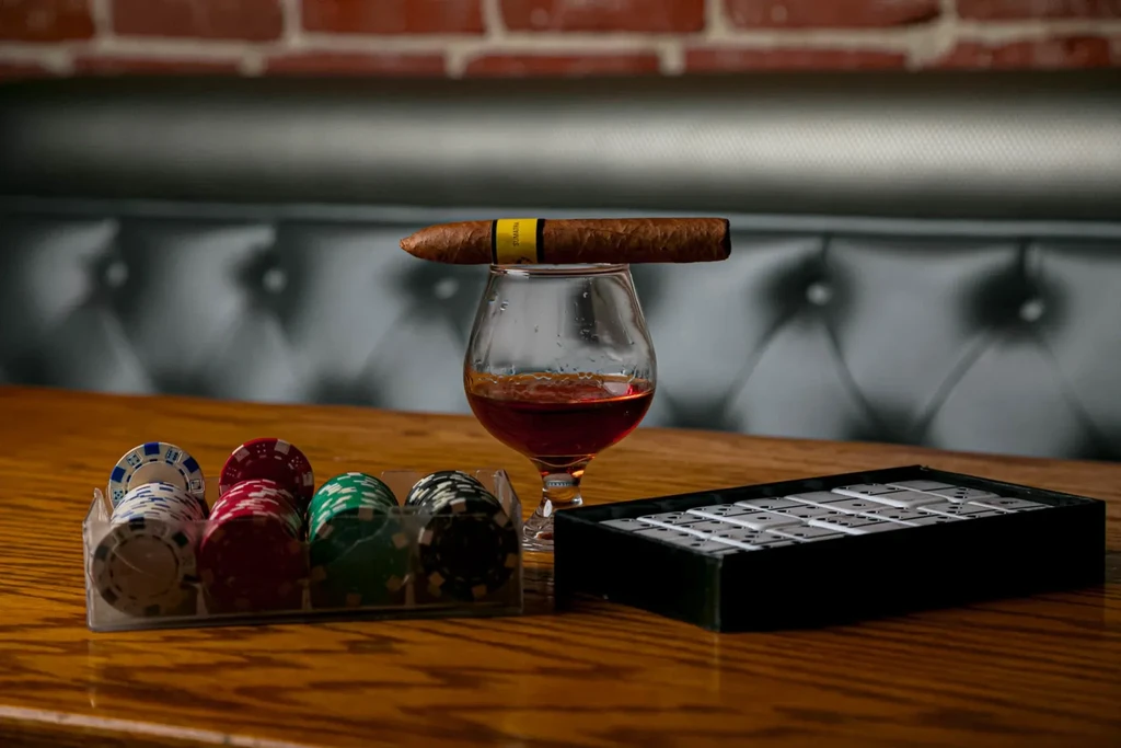 Snifter with cigar, poker chips, and dominoes on a bar table.