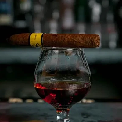 Brown cigar with yellow band on a snifter of amber spirit.