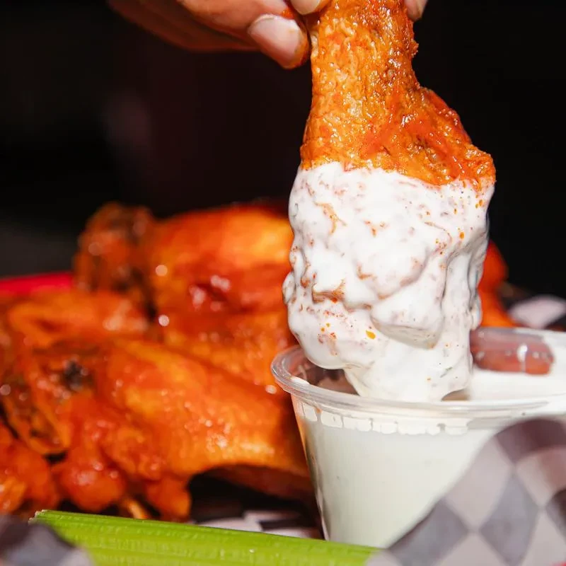 Hand dipping an orange-red buffalo wing into creamy white sauce.