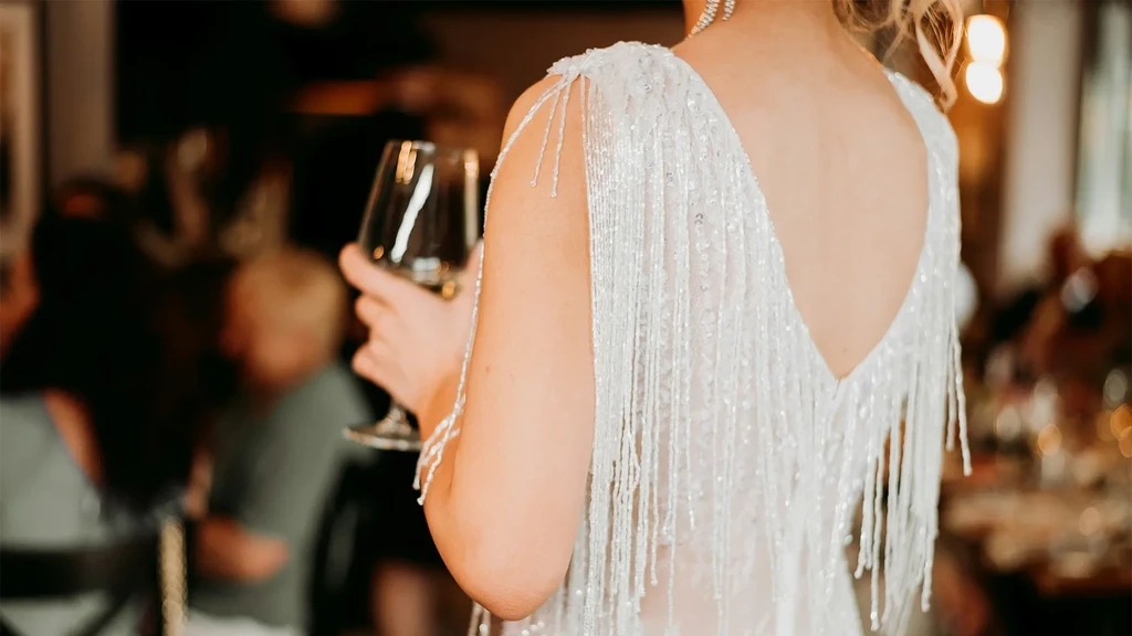 Over-the-shoulder shot: woman in white beaded dress holds wine glass.