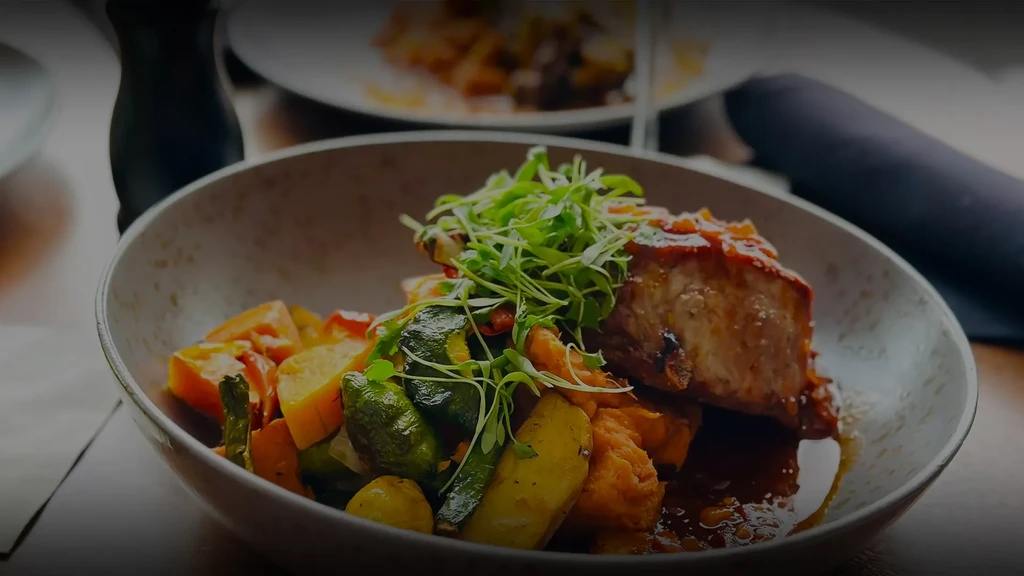 Glazed protein, microgreens, vegetables, and dark sauce in ceramic bowl.