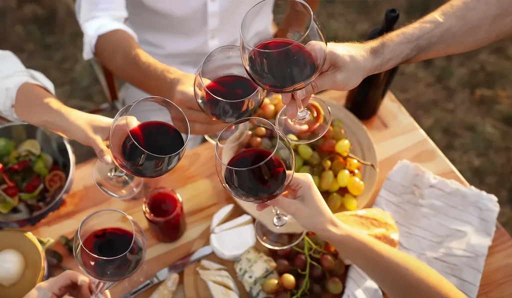 Hands toasting red wine at a rustic outdoor table with food.