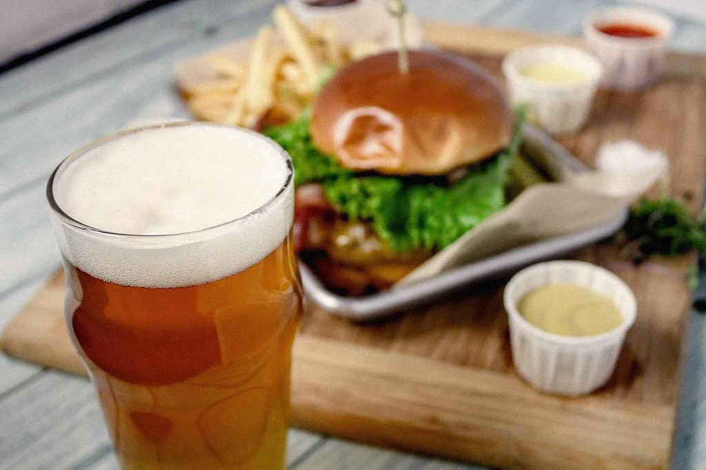 Close-up golden beer, blurred burger and fries meal in background.
