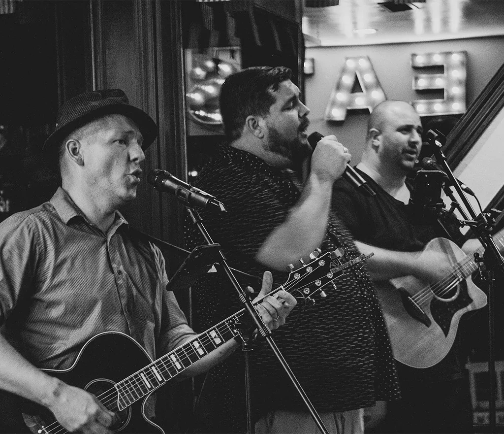 Three men perform live with guitars and microphones, black and white.