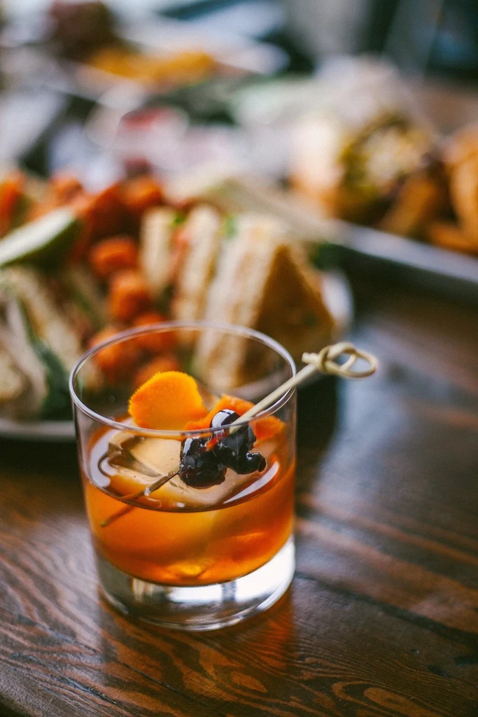 Close-up of amber cocktail, orange peel, cherries, on dark wood.