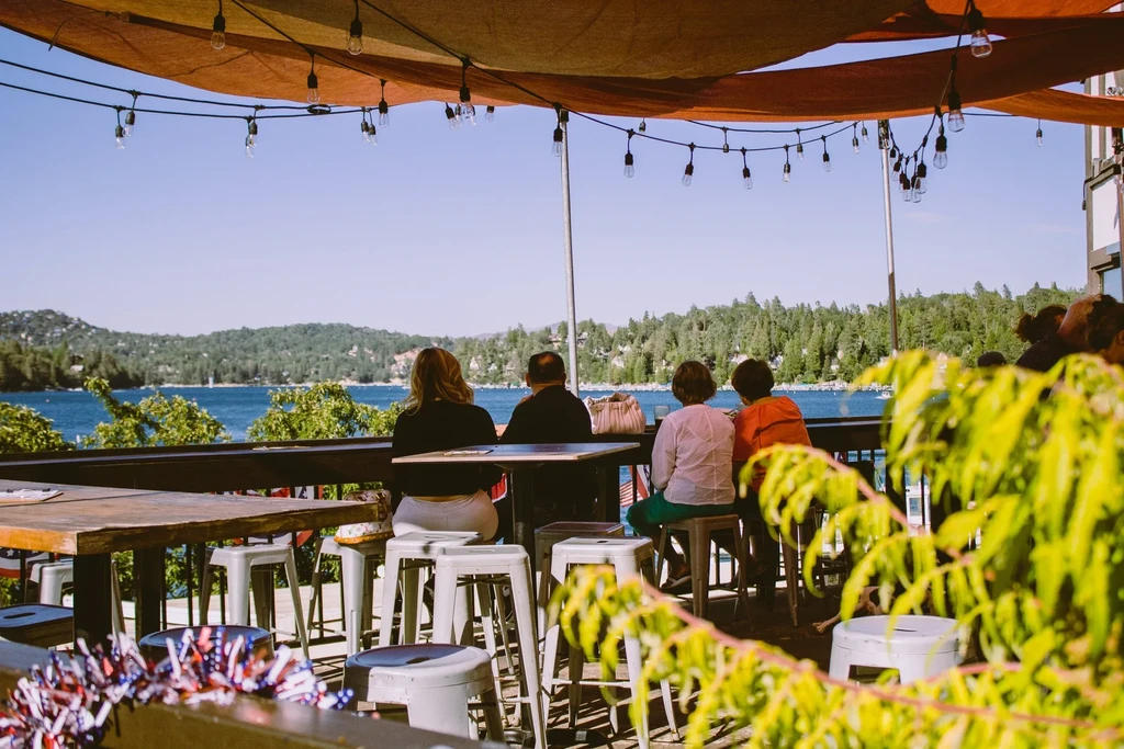 Patrons dine on a shaded patio overlooking a tranquil lake and mountains.