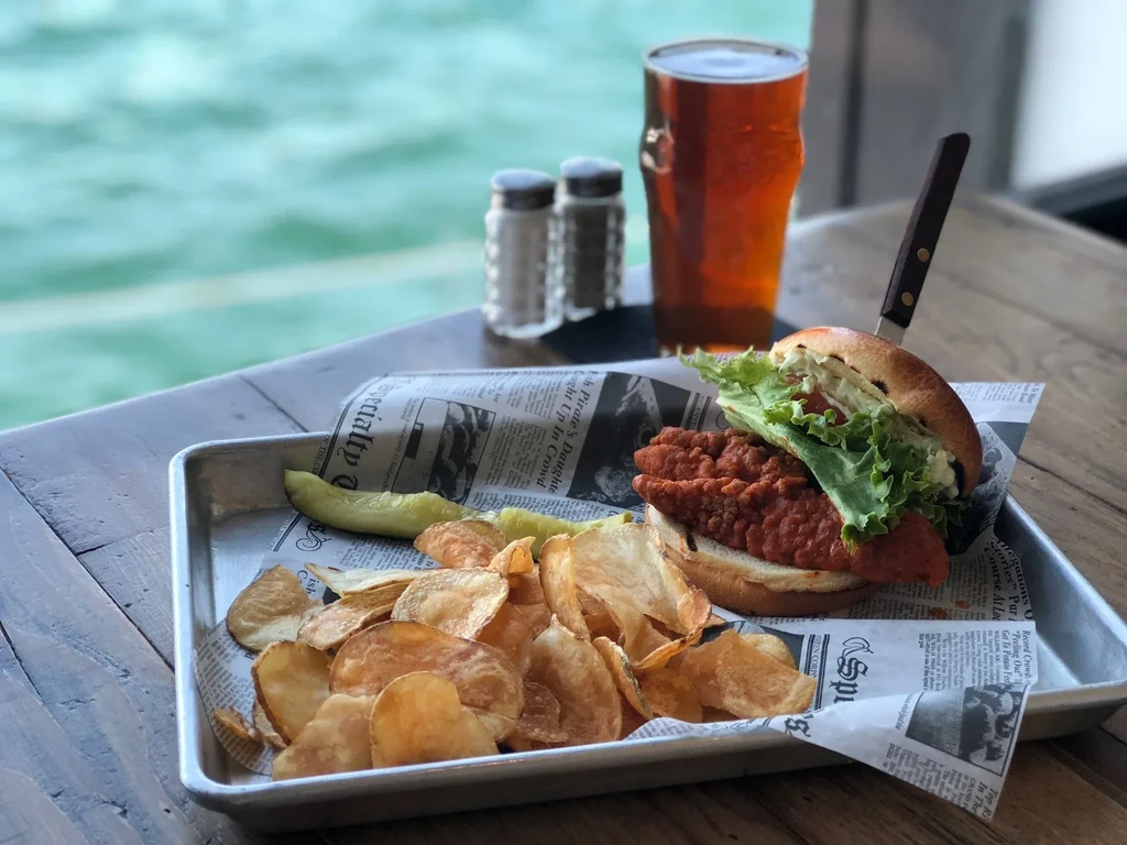 Crispy fried sandwich, chips, pickle, and drink on a waterside table.