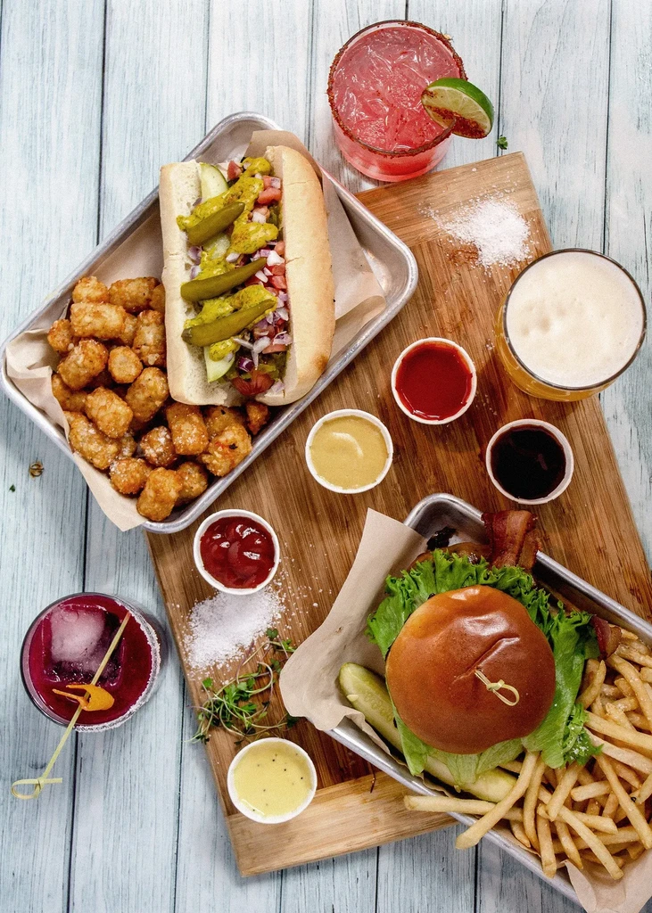 Overhead flat-lay: hot dog, burger, fries, tater tots, cocktails, beer.