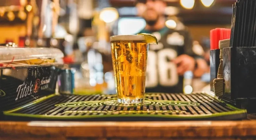 Pint of golden beer with lime on bar; blurred person visible.