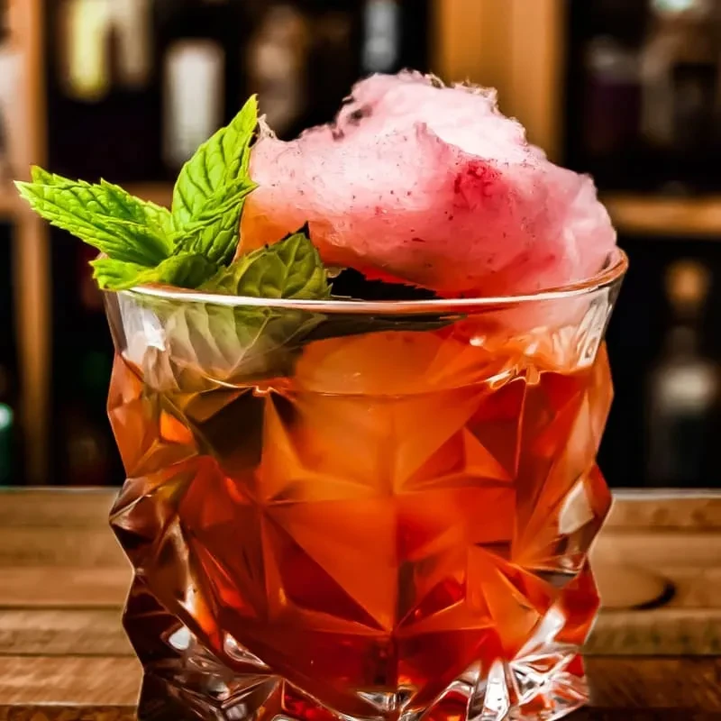 Cocktail in faceted glass, amber liquid, mint, and pink cotton candy.