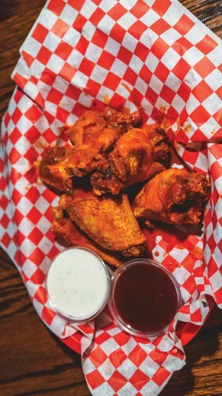 Chicken wings in red basket with checkered paper and two sauces.