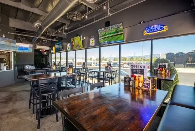 Modern industrial sports bar with high-top tables, TVs, and neon sign.