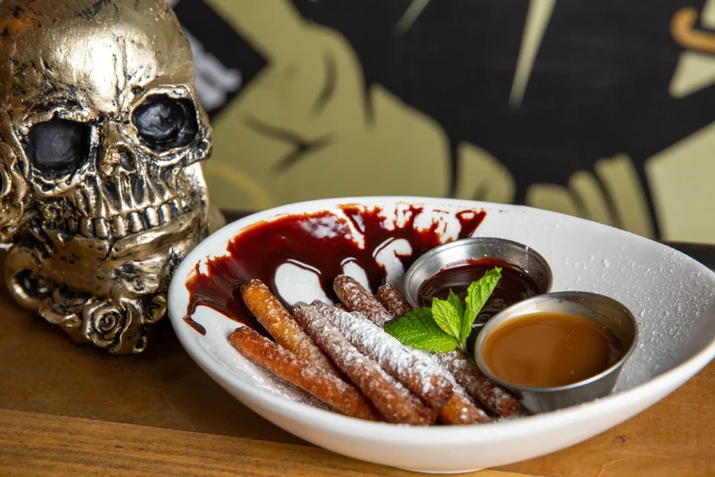 Dusted churros with chocolate and caramel sauces, beside a golden skull.