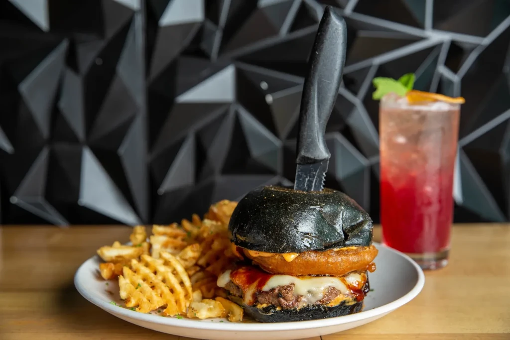 Black bun burger with onion ring, knife plunged, waffle fries, cocktail.