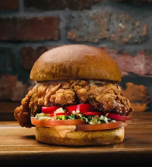 Crispy fried chicken sandwich with lettuce, tomato, sauce on wood.