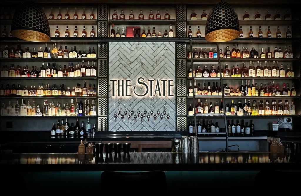 Upscale bar interior with glowing whiskey shelves and 'THE STATE' sign.