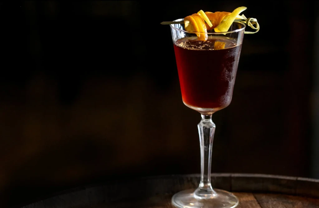 Dark cocktail in stemmed glass with citrus peel twist on wood.