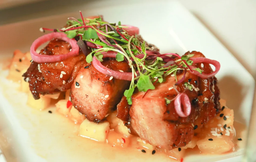 Glazed pork belly, pineapple, microgreens, and pickled onions on a plate.