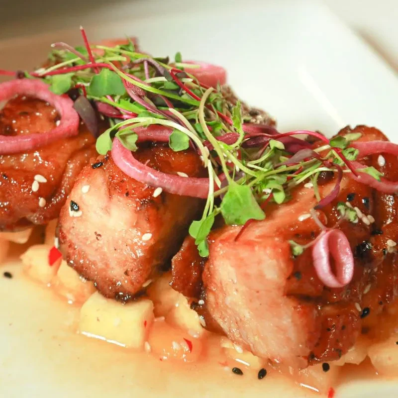 Glazed pork belly, microgreens, pickled onion, and diced fruit on white plate.