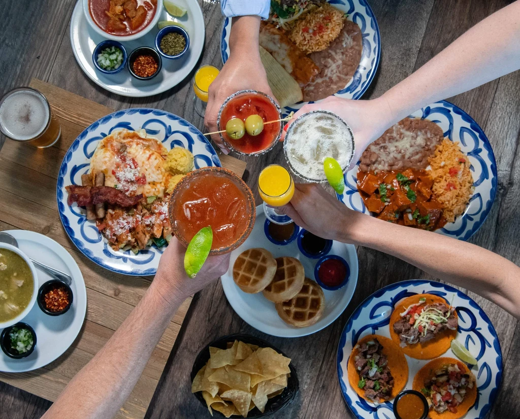 Overhead: Mexican food, drinks, and hands toasting on a wooden table.