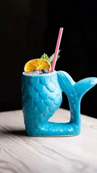 Mermaid tail mug cocktail garnished with orange, blueberries, mint, pink straw.