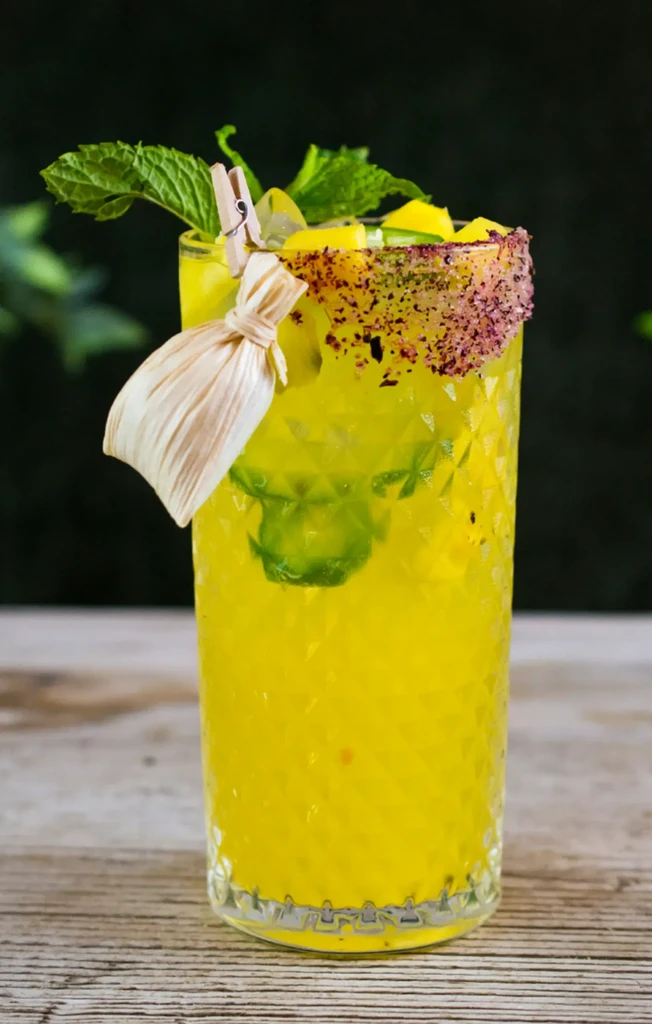 Yellow cocktail in diamond-patterned glass with chili-lime rim, corn husk clip.