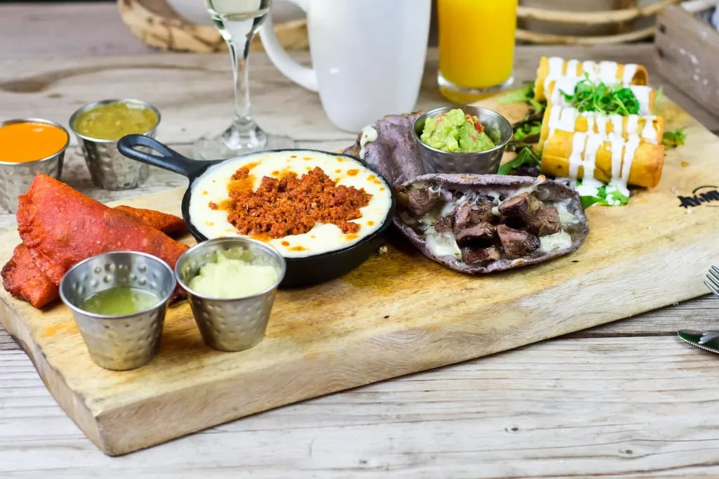 Brunch platter with skillet cheese, purple tortillas, rolled items, sauces, and drinks.