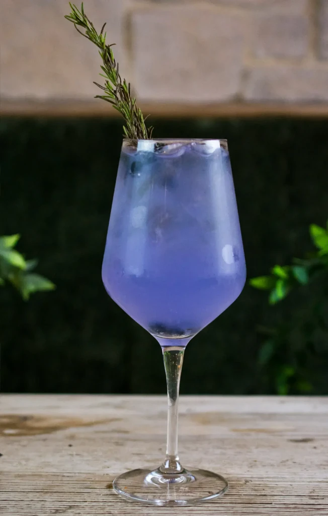Vibrant purple drink with rosemary and ice in stemmed glass.