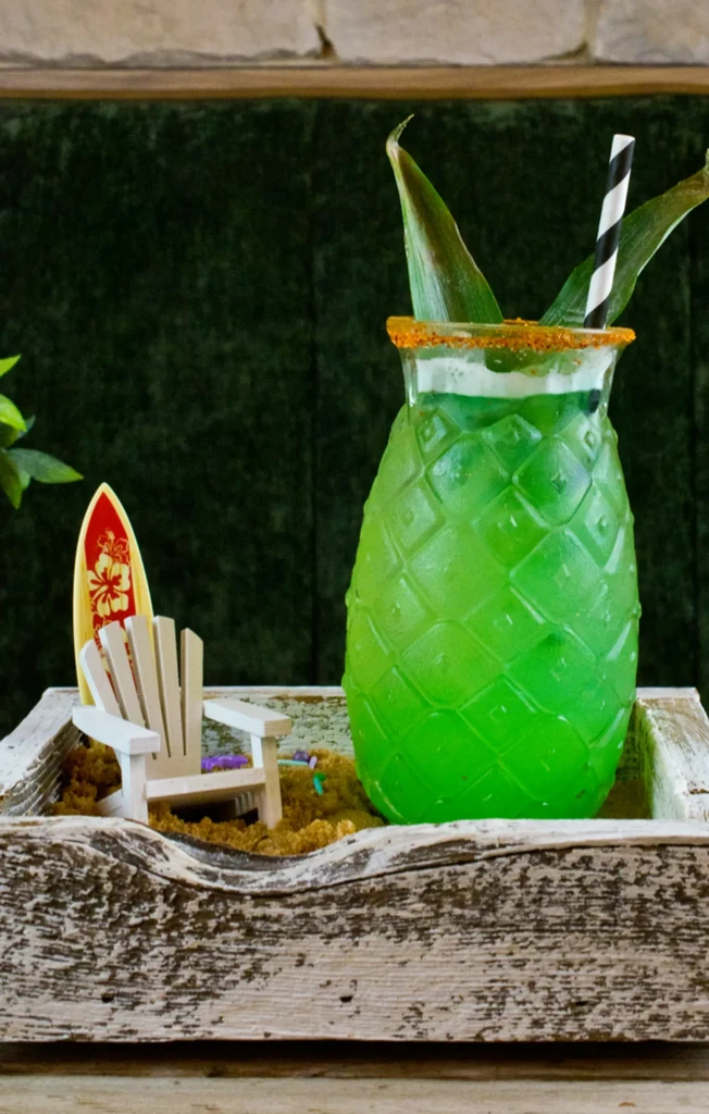 Green tropical drink in pineapple glass on a miniature beach tray.