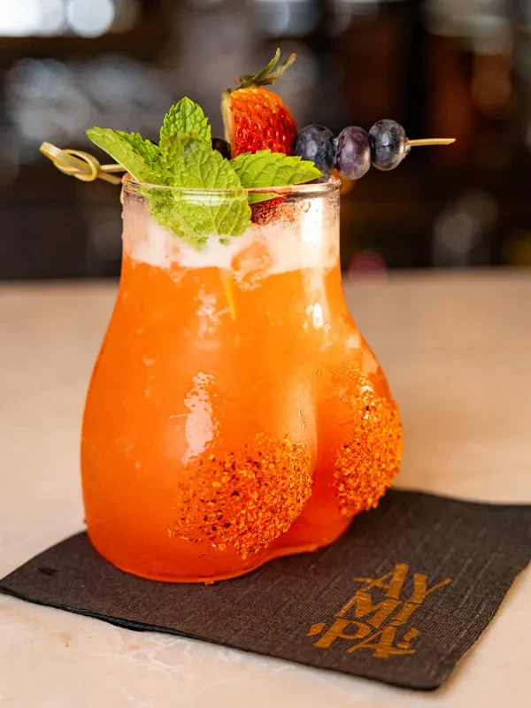 Orange cocktail in buttocks-shaped glass, garnished with mint, strawberry, blueberries.