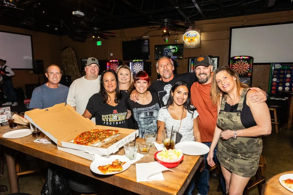 Ten adults smile, eat pizza and drink at a casual bar.