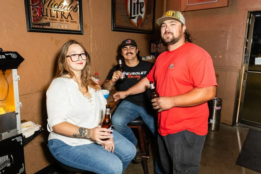 Woman and two men holding beers in a bar, looking at camera.