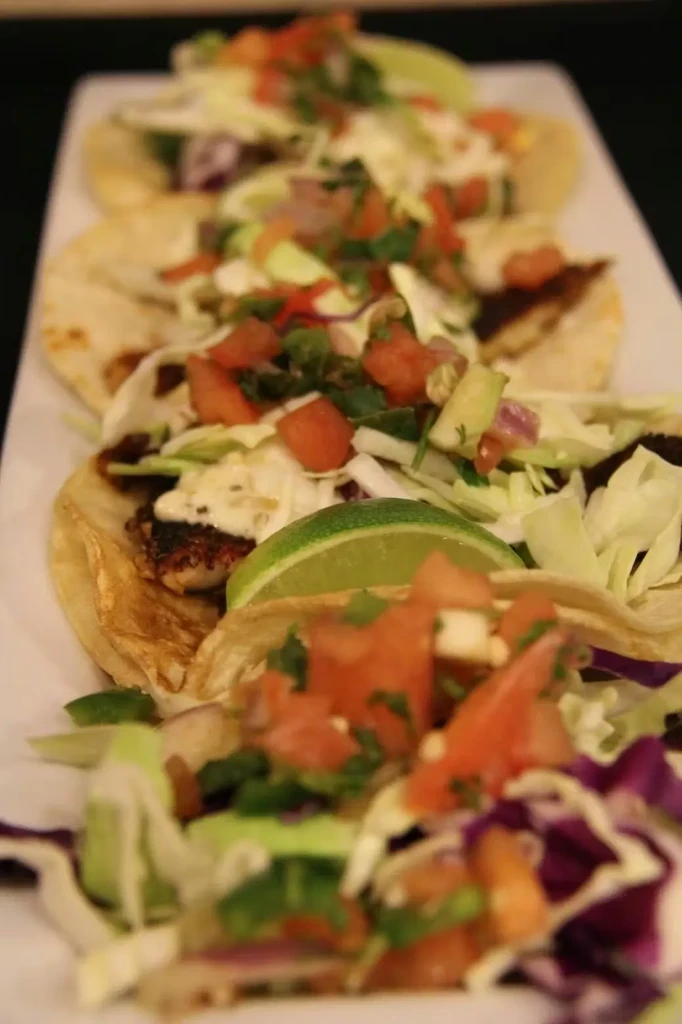 Five soft tacos with fish/meat, vegetables, and lime on white plate.